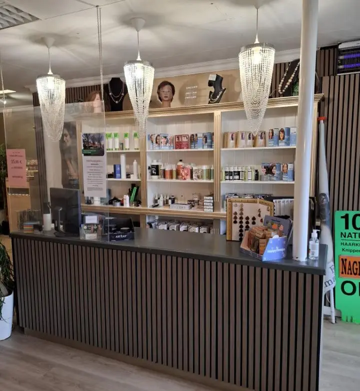 Receptiebalie van een kapsalon met verticale houten lattenpanelen voor, drie kristallen hanglampen erboven, een plexiglas scherm en een achterwand met planken vol haarverzorgingsproducten en mannequin­hoofden