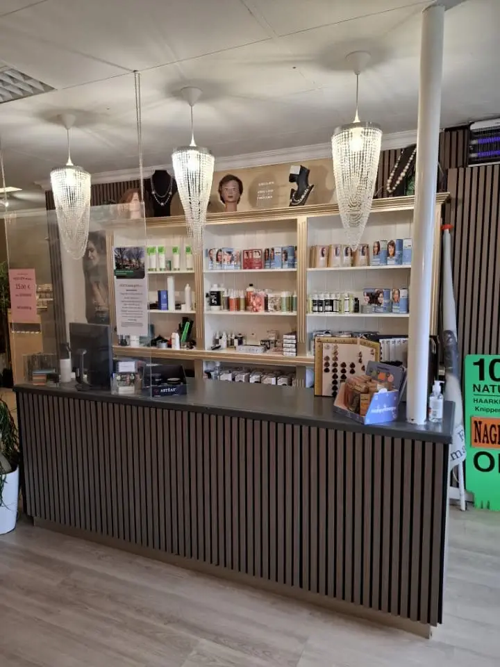 Receptiebalie van een kapsalon met verticale houten lattenpanelen voor, drie kristallen hanglampen erboven, een plexiglas scherm en een achterwand met planken vol haarverzorgingsproducten en mannequin­hoofden